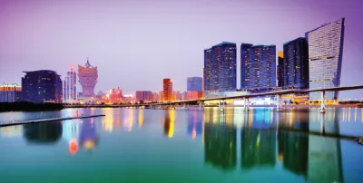 A colorful photo of the Macau skyline at dusk, with modern buildings and the iconic Grand Lisboa hotel reflected in the water.
